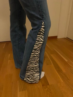 Blå bootcut jeans med zebramönstrade detaljer - Unika blå jeans med utsvängda ben och zebramönstrade kilar längs sidorna. Jeansen har klassisk femficksmodell och normal midja. Perfekta för dig som vill sticka ut med en cool och trendig look. Midjemåttet är 39 och innerbenslängden 80💓