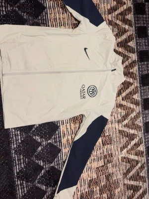 Nike Inter Milan vindjacka beige - Snygg beige vindjacka från Nike med Inter Milan-logga och Qatar Airways-tryck på bröstet. Jackan har hel dragkedja, ståkrage och mörkblå detaljer längs ärmarna. Perfekt för dig som gillar sportig stil och vill representera ditt favoritlag.