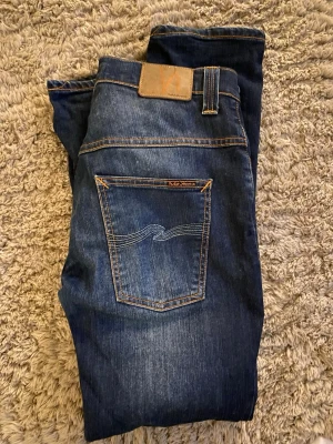 Mörkblå jeans från Nudie Jeans - Snygga mörkblå jeans från Nudie Jeans med klassisk femficksdesign och kontrastsömmar. Bakfickan har den ikoniska vågade sömmen och en liten logotyp. Perfekta för en avslappnad och trendig look.