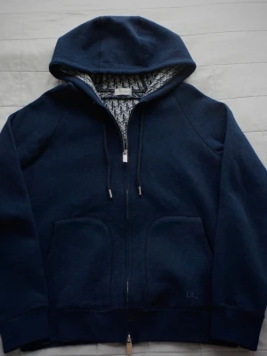 Mörkblå Designer Cardigan   - Snygg mörkblå designer cardigan med ingraverad dragkedja, huva och dragsnören. Insidan av huvan har ett mönstrat monogram-tyg i vitt och blått. Hoodien har två stora fickor framtill och diskret broderad logga vid ena fickan. Tillverkad i mjukt med väldigt tjockt bomullsmaterial. Hör av er vid minsta funderingen! 