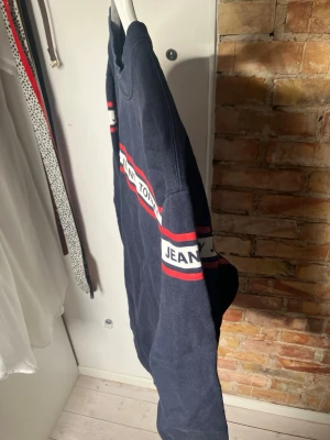 Mörkblå sweatshirt Tommy Jeans - Mörkblå sweatshirt från Tommy Jeans med vita och röda ränder över bröstet och armarna där det står 'TOMMY JEANS'. Tröjan har rund halsringning och ribbade muddar vid ärmslut och nederkant. Perfekt för en avslappnad och sportig stil.