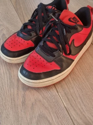 Nike röda och svarta sneakers - Snygga sneakers från Nike i rött och svart med klassisk swoosh på sidan. Skorna har svarta skosnören, perforerad tåbox och vit mellansula med röd yttersula. Tillverkade i läder och textil för en sportig look.. En liten slitning på ena skon. Men inget som förstör komforten. 
