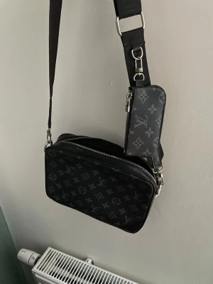 Louis Vuitton svart axelväska monogram - Säljer en svart axelväska från Louis Vuitton med klassiskt monogrammönster i grått. Väskan har justerbar axelrem, silverfärgade detaljer och en liten avtagbar ficka. Tillverkad i slitstarkt läderliknande material. Perfekt för dig som vill ha en stilren och lyxig look. AA KOPIA