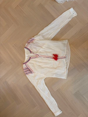 Krämvit blus med röda detaljer - Krämvit blus i tunn, luftig bomull med långa ärmar och knappar vid ärmslut. Blusen har blåa och rosa ränder på axlarna samt dekorativa rosa tofsar vid halsringningen. Perfekt för en avslappnad men trendig look och under varma sommardagar.