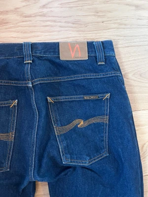 Mörkblå Nudie Jeans 32/34 - Säljer ett par mörkblå jeans från Nudie Jeans med klassisk rak passform. Jeansen har kontrastsömmar och ikoniska bakfickor med vågig söm. Tillverkade i slitstark denim av bomull. Perfekta för dig som gillar stilrena och tidlösa jeans, i princip nya använda fåtal gånger. Storlek 32/34 i fin mörkblå tvätt. Säljes då de köptes i för liten storlek.