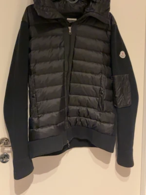 Svart dunväst med huva från Moncler - Snygg svart dunväst från Moncler med huva och dragkedja framtill. Västen har quiltad front i glansigt material, stickade ärmar och en Moncler-logga på ärmen. Praktiska fickor med dragkedja och ribbade muddar för extra komfort.