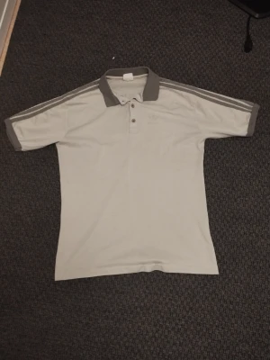 Grä adidas piké - Säljer en stilren grå pikétröja med mörkare grå krage och ärmslut. Tröjan har knappar vid halsen och ljusgråa ränder längs axlarna. Perfekt till en avslappnad look.