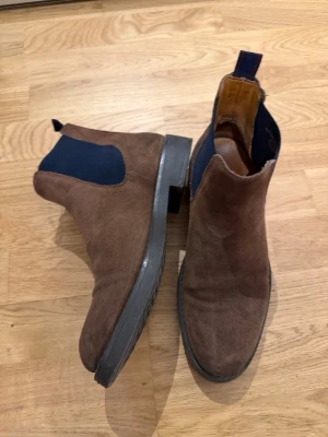 Bruna Chelsea boots från Gant - Säljer ett par bruna Chelsea boots från Gant i mocka med mörkblå resår på sidorna och robust svart sula. Klassisk siluett med dragflik bak för enkel påtagning. Perfekta för dig som vill ha en stilren och tidlös look.