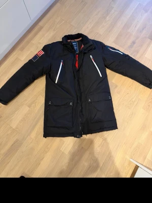SEBAGO Svart parkasjacka från sebago  - Säljer en svart varm parkasjacka från Sebago med vita och röda detaljer. Jackan har två stora fickor framtill, två snedställda bröstfickor med dragkedja och patchar på ena ärmen. Hög krage och varm vaddering, perfekt för kalla dagar.