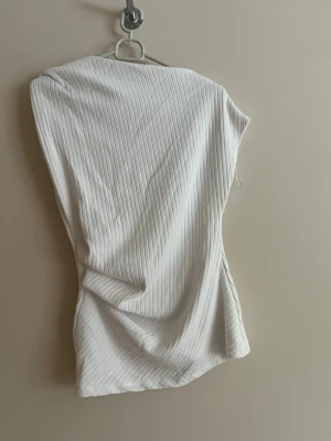 Vit ribbad topp från Zara - Säljer en stilren vit ribbad topp från Zara. Toppen har en lätt croppad passform, är ärmlös och har tunna vertikala ränder i materialet. Perfekt att matcha med jeans eller kjol för en clean look.
