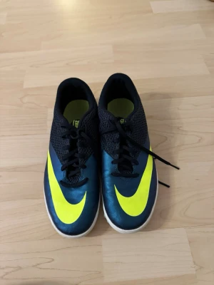 Nike blå-gula fotbollsskor - Snygga fotbollsskor från Nike i mörkblått med stora gula swoosh-loggor på ovansidan och sulan. Skorna har svart snörning och vit yttersula med hexagonmönster för bra grepp på planen. Perfekta för dig som vill sticka ut på fotbollsplanen.