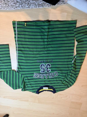 Grön randig tröja från FIX - Grön långärmad tröja från FIX med mörkblå horisontella ränder och tryck på bröstet med texten 'COLLEGE 35'. Rund halsringning med blå och gul kant. Perfekt för en avslappnad och sportig stil.