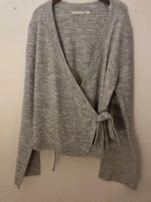 Grå omlottkofta i mjuk stickning - Säljer en ljusgrå omlottkofta i mjuk stickad kvalitet. Koftan har långa ärmar, ribbade kanter och knyts i sidan med ett band. Perfekt för lager på lager och enkel att styla till olika outfits.
