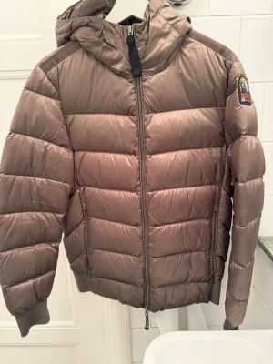 Brun/grön dunjacka Parajumpers Sheen - Snygg brun dunjacka från Parajumpers, modell Sheen. Jackan har huva, gul insida och klassisk patch på ärmen. Quiltad design med dragkedja framtill och två sidofickor. Perfekt för kalla vinterdagar och riktigt varm tack vare dunfyllningen.