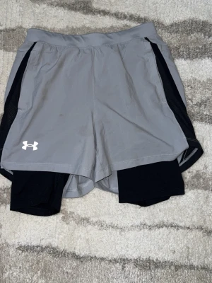 Grå träningsshorts från Under Armour - Snygga grå träningsshorts från Under Armour med svart innerbyxa och svarta detaljer på sidorna. Shortsen har elastisk midja och är tillverkade i ett lätt och snabbtorkande material, perfekta för löpning eller gym.