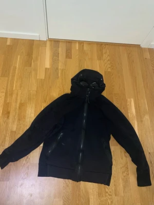Svart cp company kofta - En riktigt fin kofta från cp company som har ett par glasögon på toppen av luvan. Storlek L men passar mig utmärkt och jag är ofta en S/M och är 170 cm 53kg.