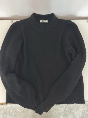 Svart stickad tröja från JDY - Snygg svart stickad tröja från JDY med puffärmar och ribbade muddar vid ärmslut och nederkant. Tröjan har en rund halsringning och är gjord i mjukt material som känns skönt mot huden. Perfekt för en trendig och enkel look.