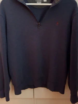 Marinblå halvzip från Polo Ralph Lauren - Klassisk marinblå tröja med halv dragkedja från Polo Ralph Lauren. Tröjan har lång ärm, ribbade muddar och en liten röd logga broderad på bröstet. Perfekt för lager på lager och enkel att matcha med olika outfits.