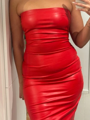 Röd axelbandslös långklänning - Säljer en snygg röd axelbandslös långklänning i figurnära modell. Klänningen har en glansig finish och är tillverkad i ett stretchigt syntetmaterial som sitter tight mot kroppen. Perfekt för dig som vill sticka ut och känna dig extra cool.
