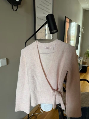 Ljusrosa omlottkofta med knytband - Supersöt ljusrosa omlottkofta med knytband i midjan och långa ärmar. Koftan har en mjuk och fluffig känsla, perfekt för att höja mysfaktorn. Enkel och stilren design som passar till det mesta.