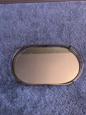 Ovalt spegelbricka med guldram - Snygg ovalt spegelbricka med ram i guldfärgad metall. Perfekt som dekoration eller för att ställa smycken, parfymer eller ljus på. Brickan har två handtag på sidorna och en stilren, modern look som passar i de flesta rum.