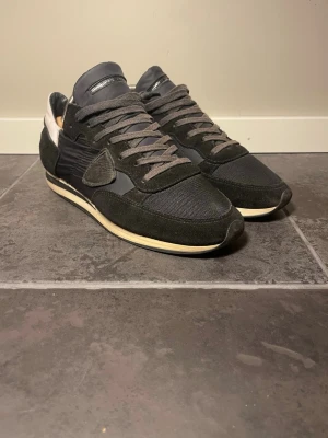 Philippe Model svarta sneakers - Säljer ett par svarta Philippe Model sneakers med klassisk låg siluett. Skorna har ovandel i mocka och mesh, detaljer i läder och en vit sula med retrovibbar. Snygga grå skosnören och diskret logga på plösen. Perfekta för dig som gillar stilrena och tidlösa sneakers.