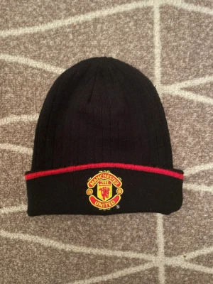 Manchester United svart mössa - Säljer en svart ribbstickad mössa från Manchester United med klubbens broderade logga framtill och en röd rand längs kanten. Perfekt för dig som vill visa ditt lagstöd även under kalla dagar.