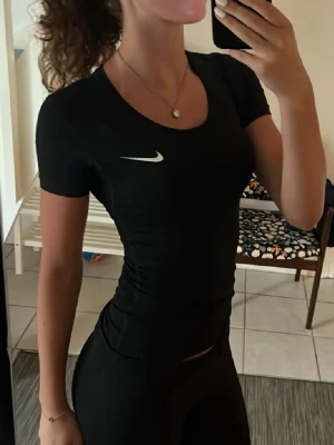 Svart Nike topp i slim fit - Säljer en svart, tight topp från Nike med korta ärmar och rund halsringning. Toppen har en vit Nike-logga på bröstet och är gjord i ett stretchigt syntetmaterial som passar perfekt till träning eller sportiga outfits.