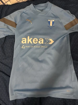 Ljusblå Malmö FF matchtröja Puma - Säljer en ljusblå Malmö FF matchtröja från Puma med klubbmärke på bröstet. Tröjan har korta ärmar och är tillverkad i ett lätt och ventilerande syntetmaterial, perfekt för fotbollsträning eller match.