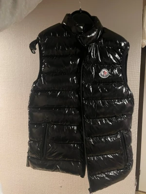 Svart glansig dunväst från Moncler - Snygg svart dunväst från Moncler med glansig finish och klassisk logga på bröstet. Västen har hög krage, två framfickor med dragkedja och quiltad design. Perfekt för lager-på-lager och ger en trendig look till din outfit. 