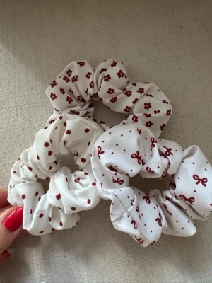 Vita scrunchies med röda mönster - Tre scrunchies i vitt tyg med olika röda mönster: blommor, prickar och rosetter. Perfekta för att piffa till håret och ge en söt touch till din look. Tillverkade i mjukt tyg som är skonsamt mot håret. Har bundlerabatt vid köp av fler varor🥰