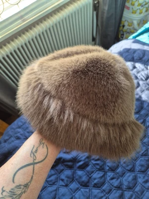 Brun vintage fuskpälsmössa - Säljer en fluffig och klassisk brun pälsmössa i rund modell. Mössan har en mjuk och tät pälsstruktur som ger en lyxig känsla och håller värmen. Perfekt accessoar för dig som vill sticka ut med retrostil och tidlös elegans.