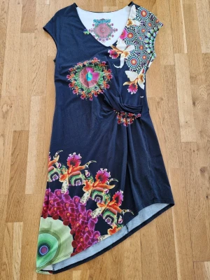 Mönstrad kortklänning från Desigual - Färgstark kortklänning från Desigual med unikt blommigt och mandalainspirerat mönster i flera färger. Klänningen har omlott-look, korta ärmar och v-ringning. Materialet är mjukt och stretchigt, perfekt för dig som gillar statement-plagg.