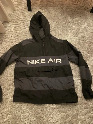 Nike Air svart/grå vindjacka med huva - Snygg vindjacka från Nike Air i svart och mörkgrått med breda blockränder. Stor logga framtill, halv dragkedja och stor ficka på magen. Jackan har huva och är tillverkad i lätt, vindtätt material. Perfekt för streetwear och sportig stil.