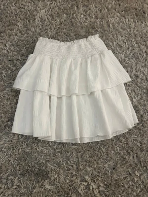 Vit volangkjol med resårmidja - Supersöt vit kortkjol med dubbla volanger och smockad resår i midjan. Kjolen har en luftig och flowy känsla, perfekt för varma dagar. Materialet känns lätt och bekvämt mot huden. Enkel att matcha med både toppar och stickade tröjor.