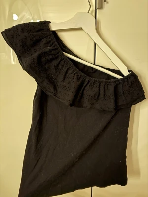 Svart oneshoulder topp med volang - Snygg svart oneshoulder topp med bred volangkant i broderad bomull. Toppen har en axel bar och volangen ger en romantisk vibe. Står varken strl eller märke men skulle gissa storlek xs/s eventuellt m