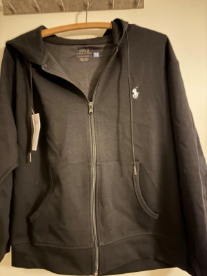 Svart hoodie från Polo Ralph Lauren - Säljer en svart hoodie från Polo Ralph Lauren med dragkedja framtill och klassisk vit logga på bröstet. Hoodien har huva med dragsko och två fickor fram. Tillverkad i mjuk bomullsblandning, perfekt för en avslappnad stil.