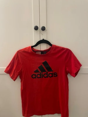 Röd Adidas t-shirt med logga - Snygg röd t-shirt från Adidas med stor svart logga på bröstet. Klassisk rund halsringning och korta ärmar. Perfekt för en sportig och avslappnad stil. Tillverkad i mjuk bomull för skön känsla hela dagen.🥰💕