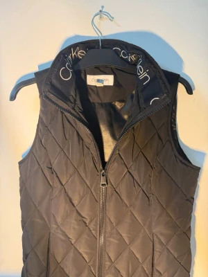 Svart quiltad dunväst Calvin Klein - Snygg svart dunväst från Calvin Klein med quiltat mönster och hög krage med logotypdetaljer. Västen har dragkedja framtill och innerficka med dragkedja. Perfekt för lager på lager och streetwear-stil.