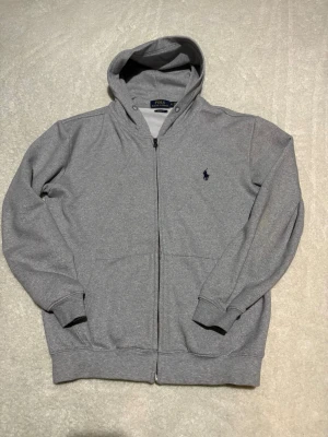 Grå zip hoodie från Polo Ralph Lauren - Snygg grå hoodie med dragkedja från Polo Ralph Lauren. Klassisk design med huva, två fickor framtill och mörkblå broderad logga på bröstet. Tillverkad i mjukt bomullsmaterial som är skönt att ha på sig. Perfekt för en avslappnad och stilren look.