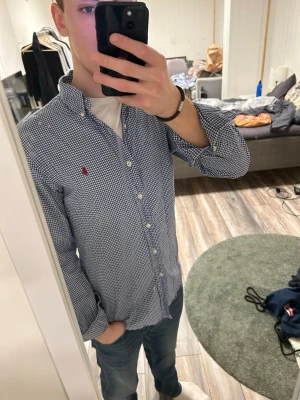 Blårutig skjorta från Ralph Lauren - Snygg blårutig skjorta från Ralph Lauren med klassisk button-down krage och broderad röd logga på bröstet. Skjortan har långa ärmar och vita knappar framtill. Perfekt till jeans eller chinos för en clean look.