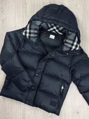 Svart dunjacka från Burberry - Snygg svart dunjacka från Burberry med klassiskt rutigt foder i huvan. Jackan har dragkedja och knappar framtill, två sidofickor med dragkedja och en vadderad, puffig design som håller dig varm. Perfekt för kalla vinterdagar och riktigt stilren.