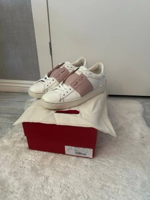 Rosa Valentinos opens  - Snygga vita sneakers från Valentino med breda ljusrosa band över vristen och diskret logga på plösen. Skorna har klassisk låg profil, rund tå och vita snören. Tillverkade i läder med detaljer i rosa och vita nyanser. Perfekta för en trendig och stilren look.