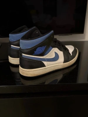 Air Jordan 1 Mid blå svart vit - Snygga Air Jordan 1 Mid sneakers i blått, svart och vitt läder. Klassisk siluett med svart snörning, blå Swoosh och ikoniska Air Jordan-loggan på sidan. Perfekta för dig som gillar streetwear och vill sticka ut med stilrena färger.