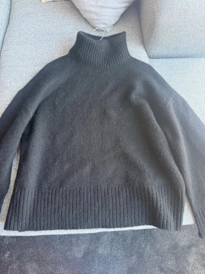 Svart polotröja från Gina Tricot - Mysig svart stickad polotröja från Gina Tricot i ullblandning. Tröjan har hög polokrage, långa ärmar och bred ribbstickad kant nedtill. Perfekt för kalla dagar och enkel att styla till olika outfits.