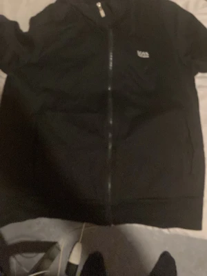 Svart zip-hoodie från Boss - Säljer en svart hoodie från boss med dragkedja framtill och liten boss-logga på bröstet. Klassisk modell med huva och fickor på sidorna. Perfekt för en avslappnad streetwear-look.