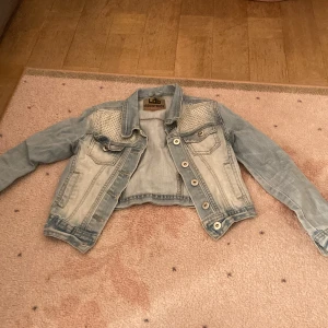 Ljusblå cropped jeansjacka Lab Industries - Snygg ljusblå jeansjacka från Lab Industries med croppad passform och coola nitar på axlarna. Klassiska bröstfickor med lock och knapp, samt dekorativa sömmar. Perfekt för dig som vill ha en trendig och unik look.