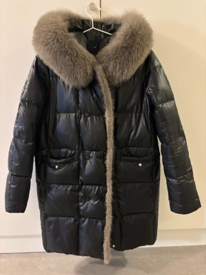 Svart lång dunjacka med pälskrage - Snygg svart dunjacka i lång modell med stor, fluffig pälskrage i gråbrunt. Jackan har två stora fickor framtill med knappar och quiltad design. Perfekt för kalla vinterdagar och ger en lyxig känsla till din outfit.