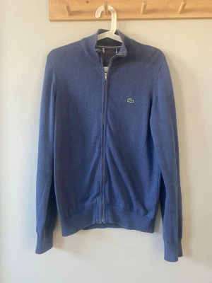 Blå ziptröja från Lacoste - Snygg blå ziptröja från Lacoste med klassisk krokodillogo på bröstet. Tröjan har ribbade muddar, hög krage och dragkedja hela vägen. Perfekt till lager-på-lager och enkel att matcha med jeans eller chinos.