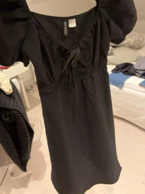 Svart kortklänning - Supersöt svart kortklänning med puffärmar, v-ringning och knyt framtill. Klänningen har en feminin siluett och är perfekt för dig som gillar en enkel men ändå trendig stil. Materialet känns mjukt och lätt.